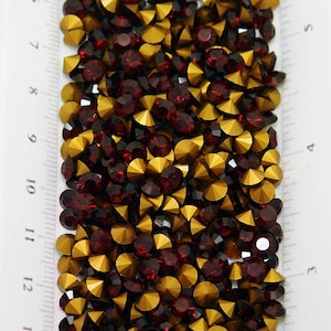 May include: Close-up of a collection of decorative rhinestones. The rhinestones are a mix of deep red and gold colors. The rhinestones are in various shapes and sizes, and are arranged in a pile. A ruler is visible on the left side.