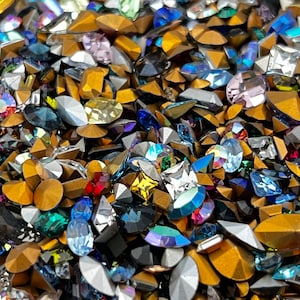 May include: A large assortment of colorful rhinestones and glass beads in various shapes and sizes. The rhinestones are mostly clear, with some having a rainbow iridescent effect. There are also some gold-colored metal pieces.