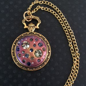 May include: A gold-tone necklace with a round pendant featuring a purple and pink iridescent background with scattered rhinestones and larger clear crystals.