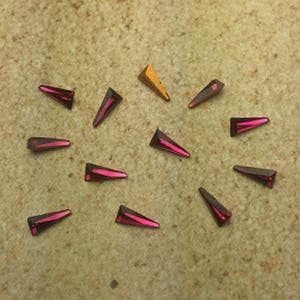 May include: A collection of 12 small, triangular rhinestones. The rhinestones are arranged in a circle on a beige surface. Most of the rhinestones are a deep pink color, while one is a gold color.
