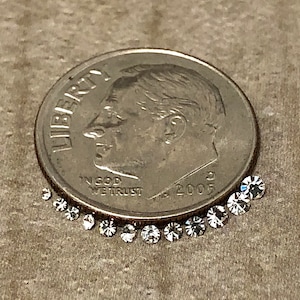 Könnte beinhalten: Eine amerikanische 10-Cent-Münze neben einer gebogenen Reihe kleiner, klarer Edelsteine. Die Münze ist silberfarben und zeigt das Profil eines Mannes. Die Edelsteine sind rund und facettiert und reflektieren das Licht. Der Hintergrund ist strukturiert.