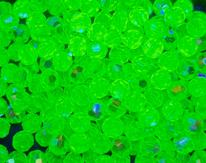 Puede incluir: Primer plano de un mont&oacute;n de cuentas facetadas de color verde ne&oacute;n. Las cuentas son redondas con m&uacute;ltiples facetas que capturan la luz, creando un efecto brillante. Las cuentas est&aacute;n dispersas, llenando el marco.
