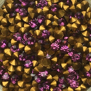 May include: A close-up image of a large quantity of small, round, gold-colored rhinestones with purple accents. The rhinestones have a flat back and a pointed top.