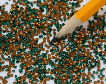 May include: A close-up image of a large quantity of green and gold rhinestones scattered on a white surface. A yellow pencil is lying on the surface, partially covered by the rhinestones.