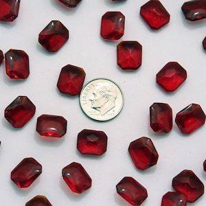 Puede incluir: Un conjunto de cuentas de vidrio rojo oscuro, facetadas y rectangulares, dispersas sobre una superficie blanca. Se incluye una moneda para comparar el tamaño. Las cuentas tienen una apariencia de gema, adecuadas para la fabricación de joyas o manualidades.