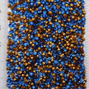 May include: A close-up image of a large quantity of blue and gold rhinestones. The rhinestones are scattered on a white surface with a ruler in the background.