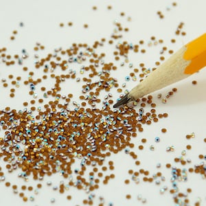 Könnte beinhalten: Nahaufnahme einer Streuung kleiner, irisierender, brauner Strasssteine. Ein gelber Bleistift mit einer angespitzen Graphitspitze berührt die Strasssteine. Die Strasssteine sind auf einer weißen Oberfläche verstreut.