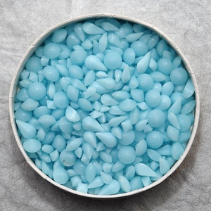 Puede incluir: Un recipiente redondo lleno de cuentas de cera azul claro de varias formas, incluyendo esferas y gotas. Las cuentas son lisas y brillantes, creando una textura visualmente atractiva. El recipiente tiene un borde blanco.