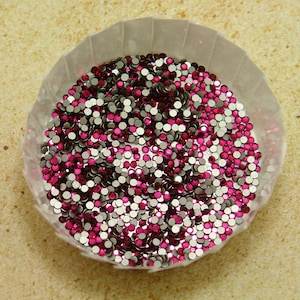 May include: A close-up shot of a small, white paper cup filled with a mix of small, round rhinestones. The rhinestones are a mix of silver, bright pink, and dark pink. The cup is sitting on a light tan surface.
