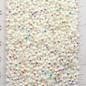 Op de afbeelding: Close-up van een stapel iriserende, gebroken witte strass-steentjes. De strass-steentjes hebben een platte achterkant en een facetgeslepen bovenkant die licht reflecteert, waardoor een regenboogeffect ontstaat. Sommige strass-steentjes hebben kleine, turquoise accenten. Een liniaal met cm-maten is zichtbaar.