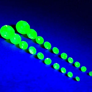 May include: A collection of 14 green gemstones of varying sizes arranged in two rows on a blue background. The gemstones are round and have a smooth, polished surface.