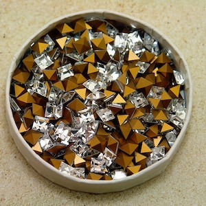 May include: A container filled with a mix of gold and clear rhinestones. The rhinestones are in various shapes, including square and pyramid.