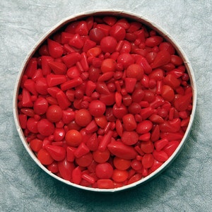 Puede incluir: Un recipiente redondo y blanco lleno de una variedad de cuentas de coral rojo brillante pulidas. Las cuentas tienen varias formas y tamaños, incluyendo esferas, lágrimas y formas irregulares. El coral tiene un acabado brillante.