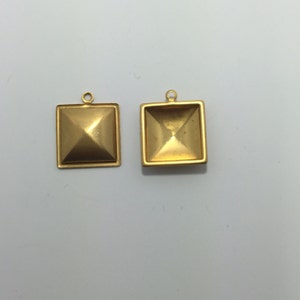 May include: Two gold-colored square pendants with pyramid-shaped centers. Each pendant has a small loop at the top for attaching to a chain or other jewelry component. The pendants are displayed against a white background.