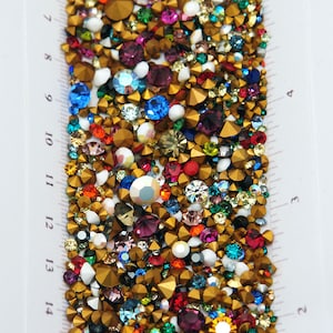 May include: A close-up of a collection of colourful rhinestones in various sizes and shapes. The rhinestones are in shades of gold, red, blue, purple, green, and white. A ruler is placed along the side, showing measurements in centimetres.