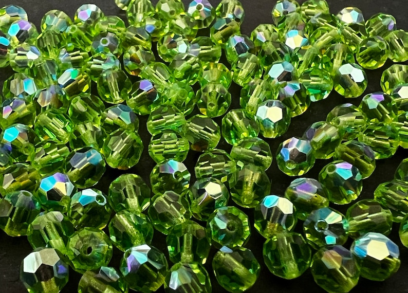 Puede incluir: Un conjunto de cuentas facetadas de color verde claro con reflejos iridiscentes. Estas peque&ntilde;as cuentas decorativas est&aacute;n dispersas, mostrando sus m&uacute;ltiples facetas y la refracci&oacute;n de la luz. Ideal para la fabricaci&oacute;n de joyas y manualidades.