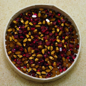 May include: A close-up of a white bowl filled with a mixture of gold and burgundy oval-shaped rhinestones.