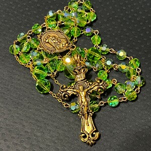 Puede incluir: Un rosario con cuentas verdes facetadas y detalles met&aacute;licos dorados. El rosario presenta un colgante de crucifijo y un medall&oacute;n con una imagen religiosa. Las cuentas est&aacute;n ensartadas en una cadena dorada.