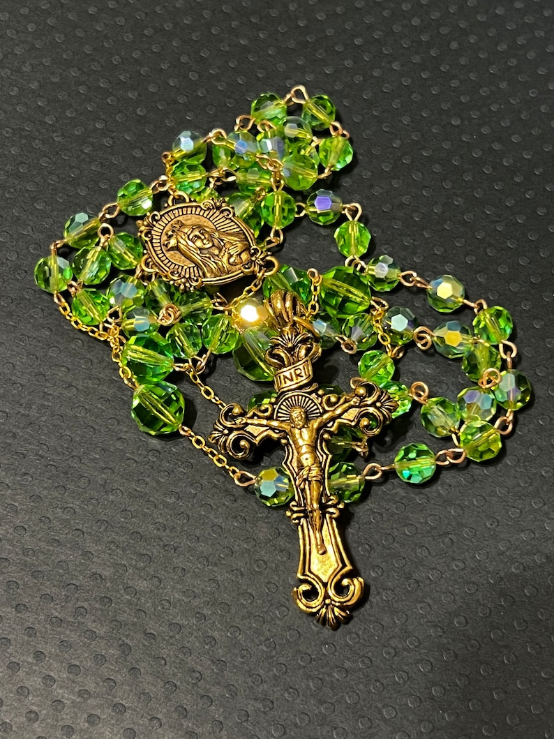 Puede incluir: Un rosario con cuentas verdes facetadas y detalles met&aacute;licos dorados. El rosario presenta un colgante de crucifijo y un medall&oacute;n con una imagen religiosa. Las cuentas est&aacute;n ensartadas en una cadena dorada.
