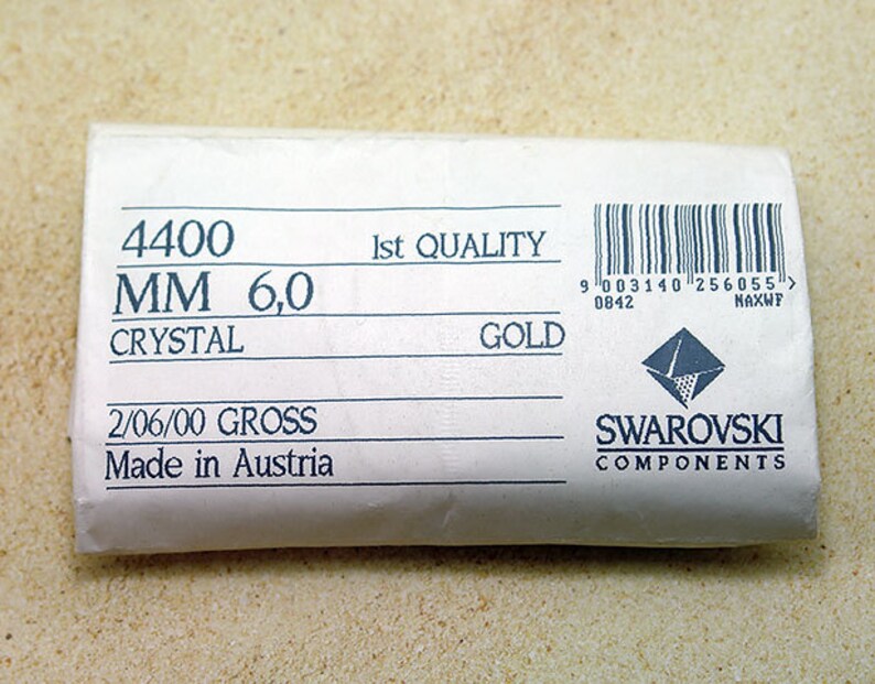 May include: A white label with black text that reads "4400 MM 6,0 CRYSTAL 2/06/00 GROSS Made in Austria 1st QUALITY GOLD" and a barcode with the text "9 003140 256055> 0842 NAXWP" and a Swarovski logo with the text "SWAROVSKI COMPONENTS".