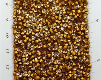 May include: Close-up of a large quantity of gold-toned, faceted, round rhinestones with a pointed back. The rhinestones are arranged in a random pattern.
