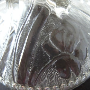 May include: A clear glass lid with a textured design featuring a flower pattern. The lid has a raised edge and a smooth, polished surface.