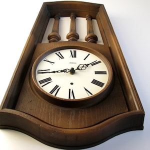 Puede incluir: Un reloj de pared de madera vintage con esfera blanca y números romanos negros. El reloj tiene un marco de madera marrón oscuro y husillos decorativos en la parte superior. Las manecillas del reloj son negras y la esfera dice "Sunbeam".