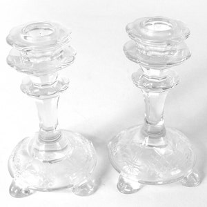 May include: Two clear glass candle holders with a scalloped design and three feet. The candle holders are made of crystal and have a delicate, elegant look.