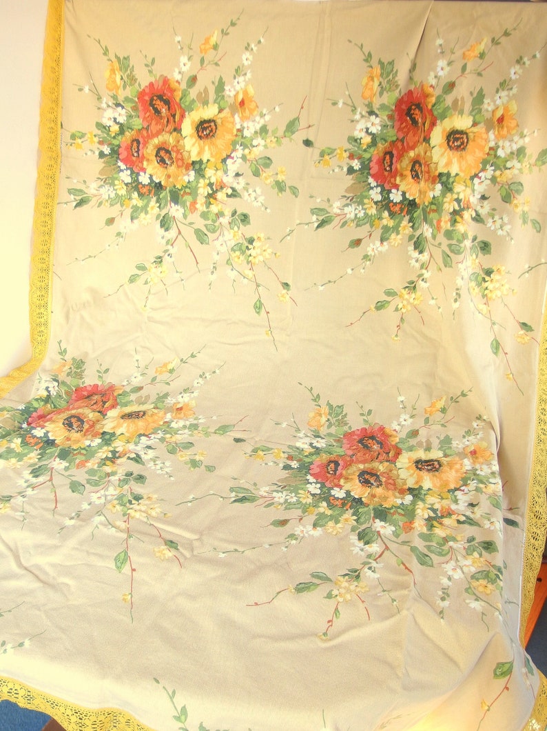 May include: A beige tablecloth with a yellow lace border features a floral pattern of yellow, orange, and red flowers. The flowers are arranged in bouquets and are scattered across the tablecloth.