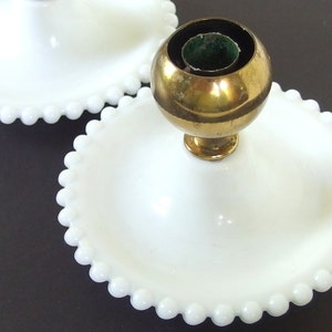 May include: Two white milk glass candle holders with a beaded edge. The candle holder on the right has a brass candle cup.