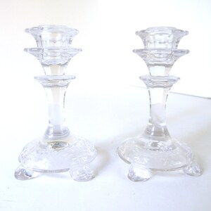 May include: Two clear glass candle holders with a tiered design and a base with a decorative pattern. The candle holders have three legs each.