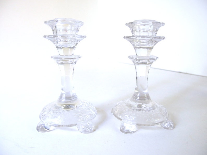 May include: Two clear glass candle holders with a tiered design and a base with a decorative pattern. The candle holders have three legs each.