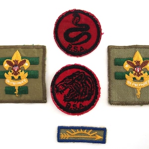 May include: A collection of vintage Boy Scout patches. Includes two square patches with a gold fleur-de-lis emblem, two round red patches with black animal designs, and a blue rectangular patch with a yellow arrow and sun design.