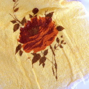 May include: A yellow bath towel with a brown and orange floral design. The flower is a rose with a stem and leaves.
