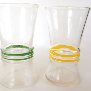 May include: Two clear glass tumblers with a flared top and base. One tumbler features three horizontal green rings around the center, while the other tumbler features three horizontal yellow rings around the center of the glass body.
