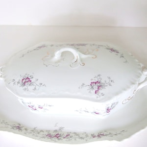 Puede incluir: Plato de servir de porcelana blanca con tapa. El plato tiene un borde festoneado y está decorado con un patrón floral en tonos rosa y gris. La tapa tiene una manija decorativa.