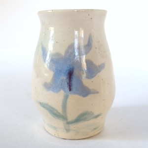 May include: A small, white ceramic vase with a blue flower painted on the front. The vase has a rounded body and a slightly flared opening.