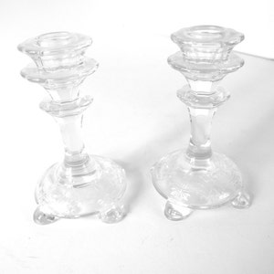 May include: Two clear glass candle holders with a decorative design on the base. Each holder has three legs and a tiered top.
