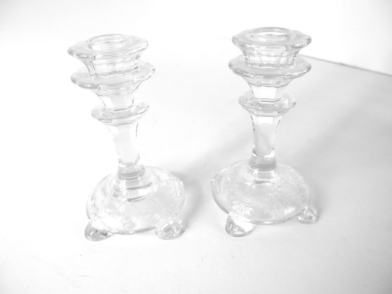 May include: Two clear glass candle holders with a decorative design on the base. Each holder has three legs and a tiered top.