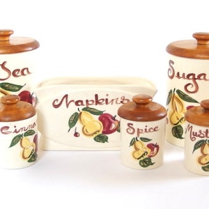 Puede incluir: Conjunto de seis botes de cocina de cerámica vintage con tapas de madera. Los botes están decorados con un motivo de peras y manzanas y tienen etiquetas escritas a mano: "Sea", "Cinnamon", "Napkins", "Spice", "Sugar" y "Mustard".