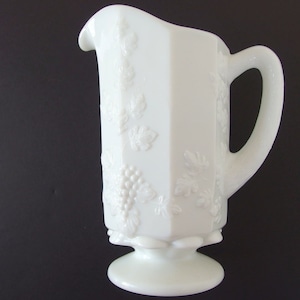 May include: White milk glass pitcher with a grapevine design. The pitcher has a footed base and a handle.