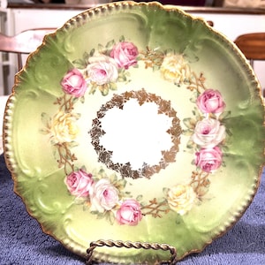 Puede incluir: Un plato decorativo vintage con borde festoneado, con fondo verde y crema. El plato está adornado con un anillo de rosas rosas y amarillas y detalles dorados. Se exhibe sobre un soporte de metal retorcido.