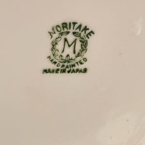 Puede incluir: Primer plano de una superficie de cerámica blanca con un logotipo verde de Noritake. El logotipo incluye la palabra "NORITAKE" sobre una "M" estilizada dentro de una corona, con "HAND PAINTED" y "MADE IN JAPAN" debajo.