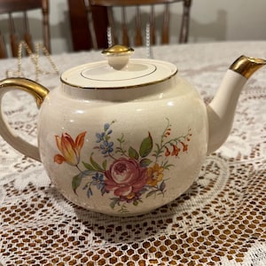 May include: Cream-colored ceramic teapot with a floral design featuring pink roses, orange tulips, and blue flowers. The teapot has a gold-colored handle, spout, and lid finial. The teapot is sitting on a white lace tablecloth.