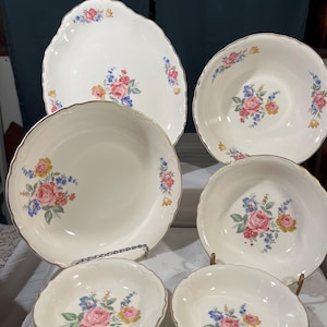 May include: A collection of white porcelain serving dishes with a floral pattern. The set includes a large platter, bowls, and smaller dishes. Each piece features pink roses, blue and yellow flowers, and gold trim.