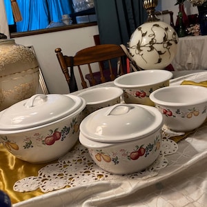 May include: A set of white ceramic serving bowls with lids, decorated with a fruit pattern of apples, pears, and grapes. The bowls are arranged on a table with a lace doily and a gold-colored runner.