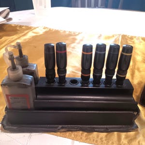 May include: A black pen holder with eight slots, holding seven black pens with colored bands. Two bottles of ink are visible in the foreground.