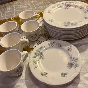 May include: A set of white ceramic plates and cups with a grape and floral design. The plates are stacked, and the cups are arranged nearby. The design includes purple grapes, green leaves, and pink flowers.