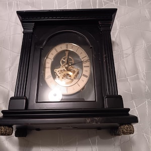 May include: A black wooden clock with a gold-toned face and exposed gears. The clock has a glass front and sits on four decorative feet.