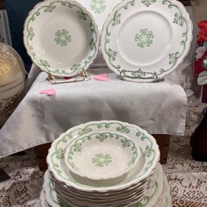 May include: A collection of white porcelain plates and bowls with a green floral design and gold trim. The plates are displayed on a white tablecloth, some on gold stands. The set includes various sizes, suitable for a formal dining setting.